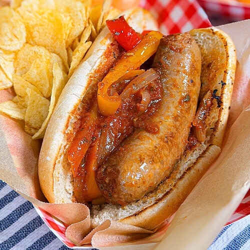 A sausage in a bun with grilled peppers and onions, served with potato chips on the side, all in a red and white paper tray on a blue and white striped cloth.