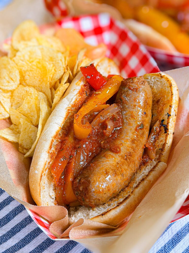 A sausage in a bun with grilled peppers and onions, served with potato chips on the side, all in a red and white paper tray on a blue and white striped cloth.