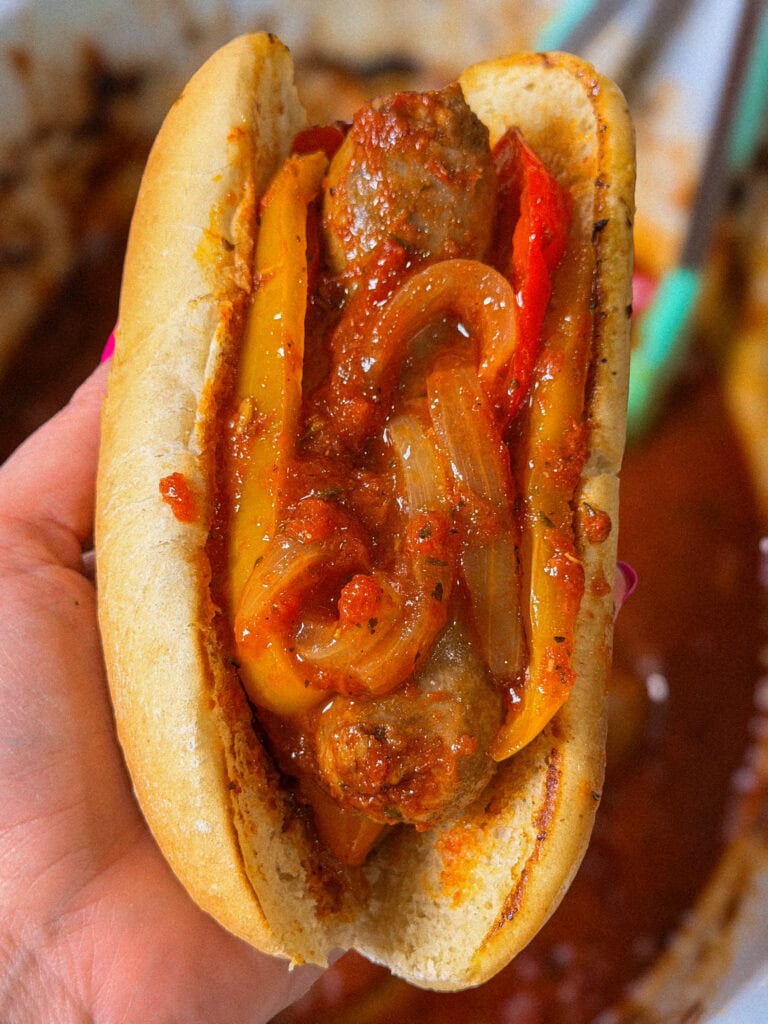 A hand holds a sausage in a toasted bun topped with sautéed onions and bell peppers in tomato sauce. Tongs and a pan of sauce are blurred in the background.