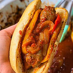 A hand holds a sausage sandwich with sautéed onions and peppers in tomato sauce, served in a toasted bun.