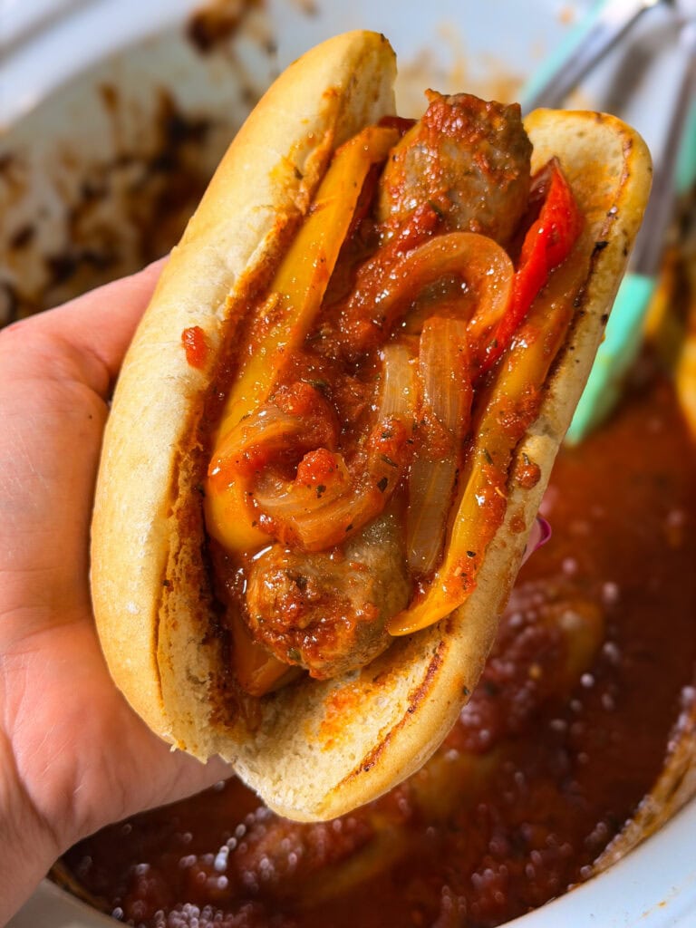 A hand holds a sausage sandwich with sautéed onions and peppers in tomato sauce, served in a toasted bun.