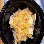 A slow cooker filled with raw chicken breasts, sauerkraut, and pats of butter, set on a blue and white striped cloth.
