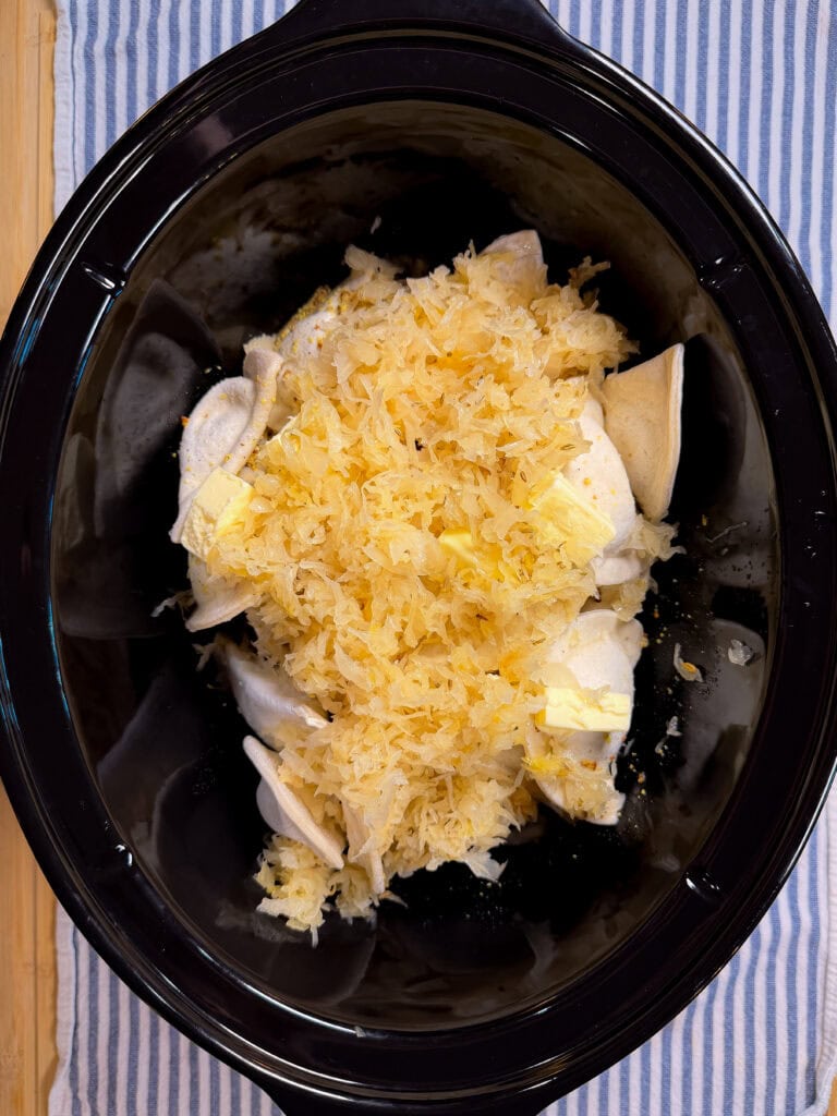A slow cooker filled with raw chicken breasts, sauerkraut, and pats of butter, set on a blue and white striped cloth.