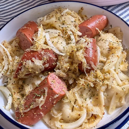 A bowl of pierogi topped with sautéed onions, sliced smoked sausage, and herbs sits on a striped cloth.