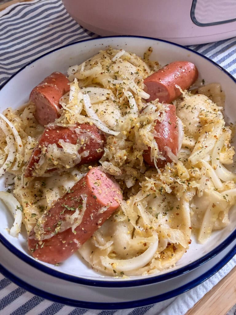 A bowl of pierogi topped with sautéed onions, sliced smoked sausage, and herbs sits on a striped cloth.
