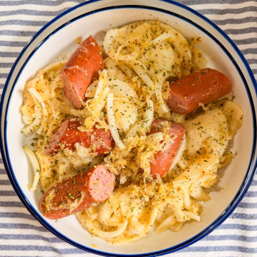 A bowl of pierogi topped with shredded cheese, sliced sausage, and herbs sits on a blue and white striped cloth.