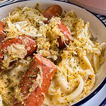A close-up of sliced sausage and onions served over pasta in a white dish, garnished with herbs and seasonings.