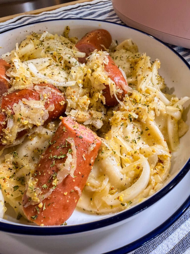 A close-up of sliced sausage and onions served over pasta in a white dish, garnished with herbs and seasonings.