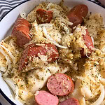 A bowl of sliced sausage mixed with pierogi, sautéed onions, and herbs, served on a white plate with a blue rim.
