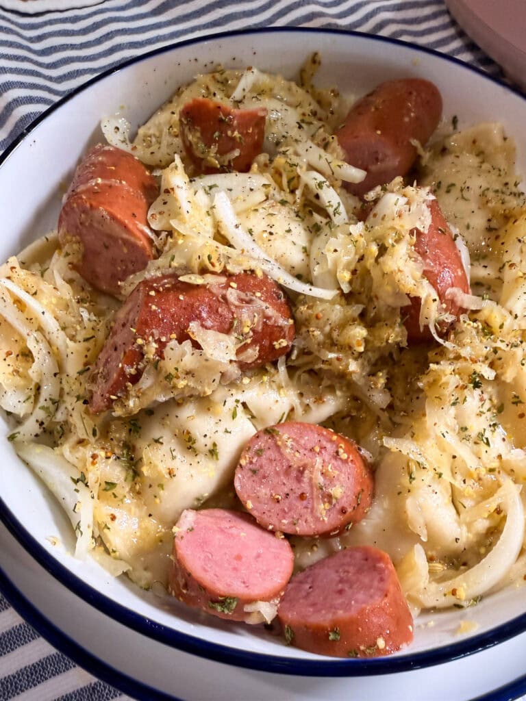 A bowl of sliced sausage mixed with pierogi, sautéed onions, and herbs, served on a white plate with a blue rim.