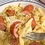 A close-up of a fork holding pierogi with sliced sausage, onions, and herbs, above a bowl filled with the same dish.