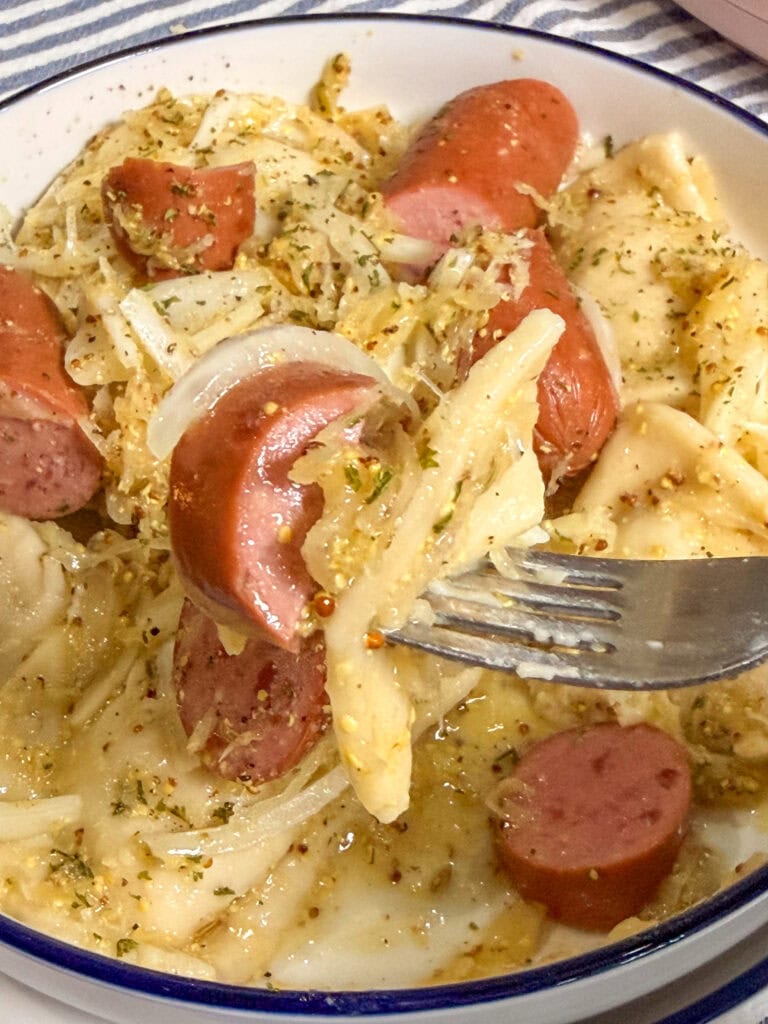 A close-up of a fork holding pierogi with sliced sausage, onions, and herbs, above a bowl filled with the same dish.