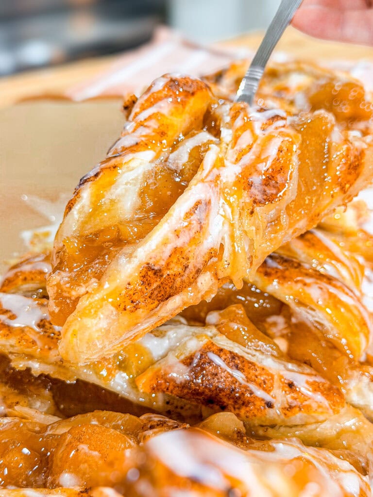 A close-up of a fork lifting a slice of apple danish pastry, topped with gooey apple filling and drizzled with white icing. The pastry looks golden brown, flaky, and sweet.