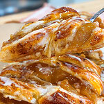 Close-up of a flaky apple pastry with visible cinnamon, gooey apple filling, and drizzle of white icing, being lifted from a stack of similar pastries.