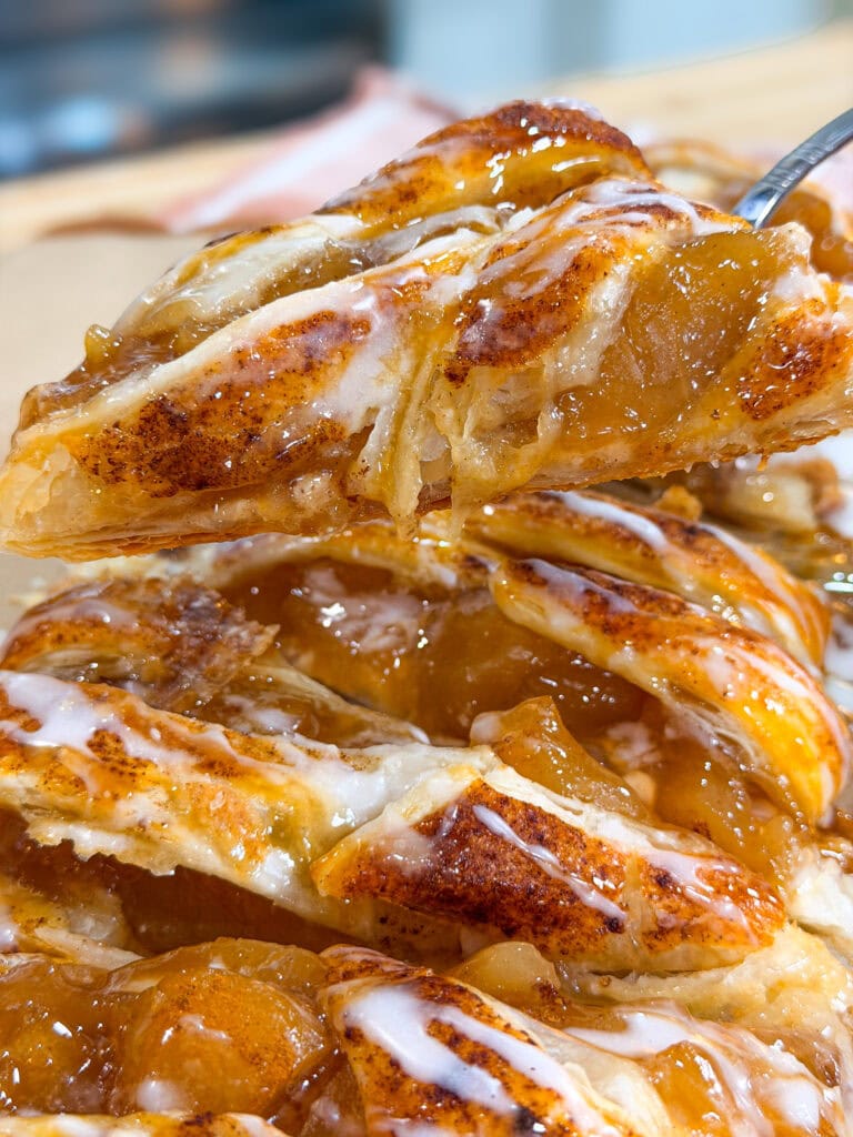 Close-up of a flaky apple pastry with visible cinnamon, gooey apple filling, and drizzle of white icing, being lifted from a stack of similar pastries.