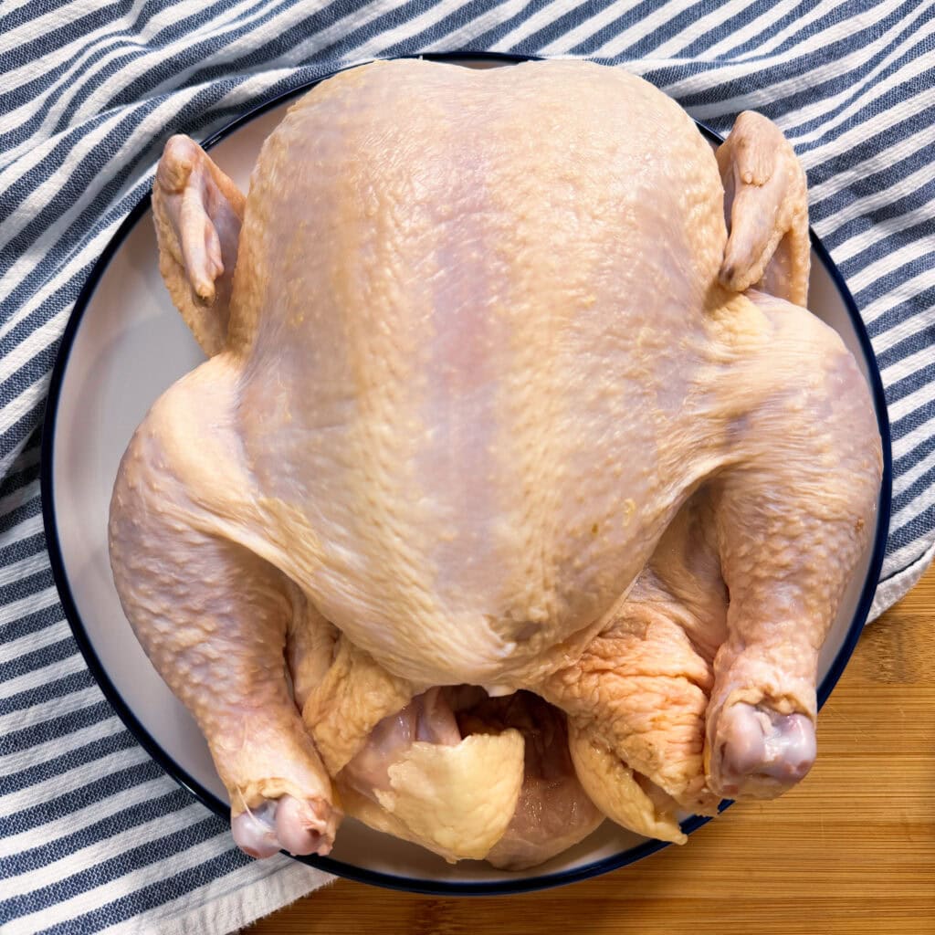 Raw whole chicken placed on a round plate, with a striped cloth in the background, ready for preparation or cooking.