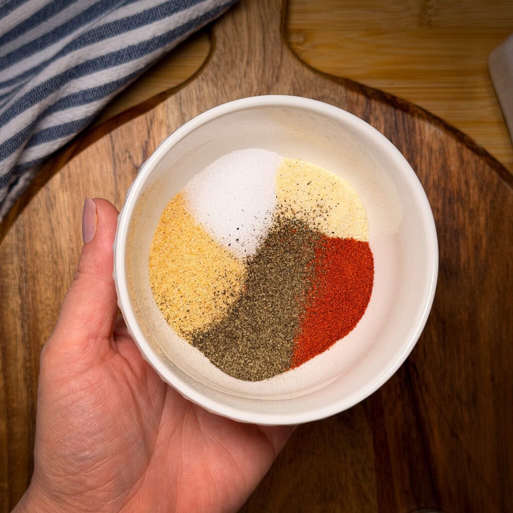 A hand holds a white bowl filled with neatly separated piles of salt, black pepper, paprika, garlic powder, and onion powder over a wooden surface.