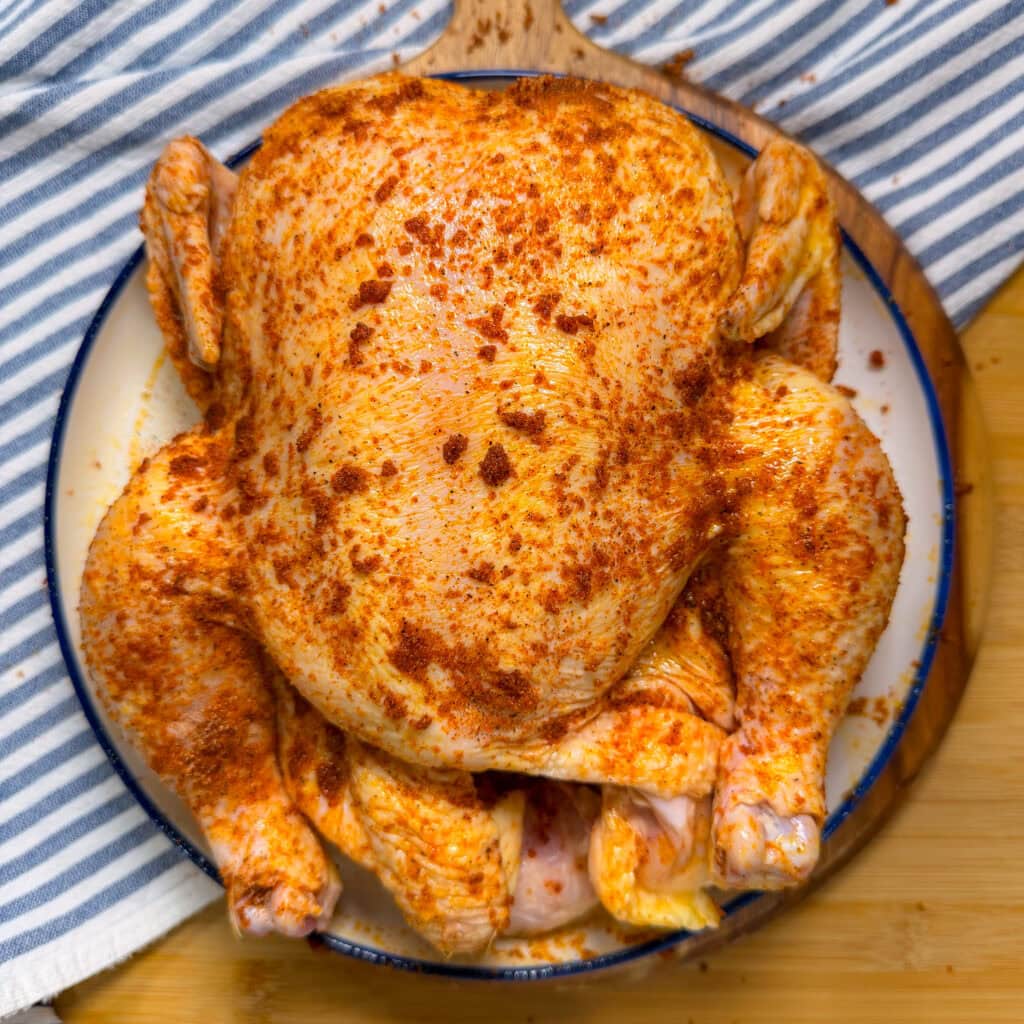 Raw whole chicken coated with red seasoning sits on a white plate with a blue rim, placed on a wooden surface next to a blue-and-white striped cloth.