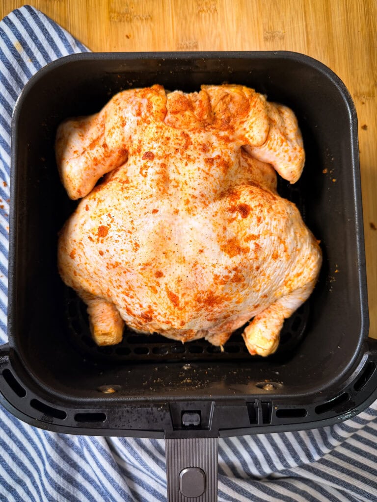 A whole raw chicken, seasoned with spices, sits in an air fryer basket. The basket rests on a blue and white striped cloth on a wooden surface.