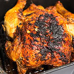 A whole roasted chicken with crispy, heavily charred skin sits in a black pan. The chicken is golden brown with dark burnt spots on top. The pan rests on a blue and white striped cloth.