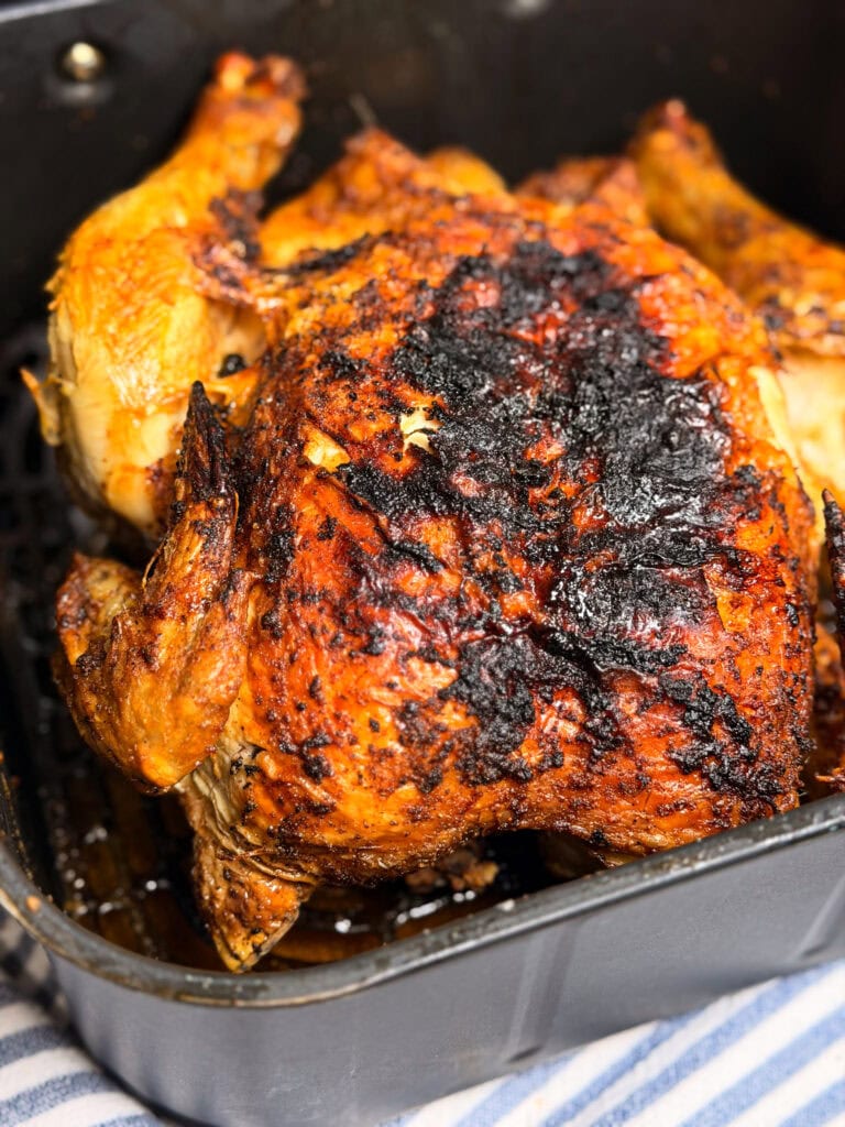 A whole roasted chicken with crispy, heavily charred skin sits in a black pan. The chicken is golden brown with dark burnt spots on top. The pan rests on a blue and white striped cloth.