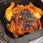 A whole roasted chicken with crispy, browned skin sits in an air fryer basket. The skin is seasoned and slightly charred in spots. The air fryer rests on a striped cloth surface.