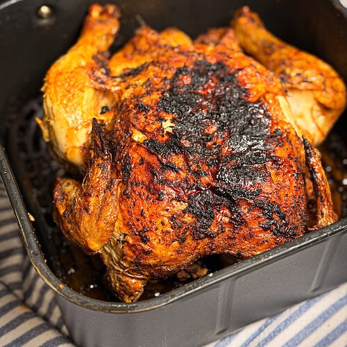 A whole roasted chicken with crispy, browned skin sits in an air fryer basket. The skin is seasoned and slightly charred in spots. The air fryer rests on a striped cloth surface.