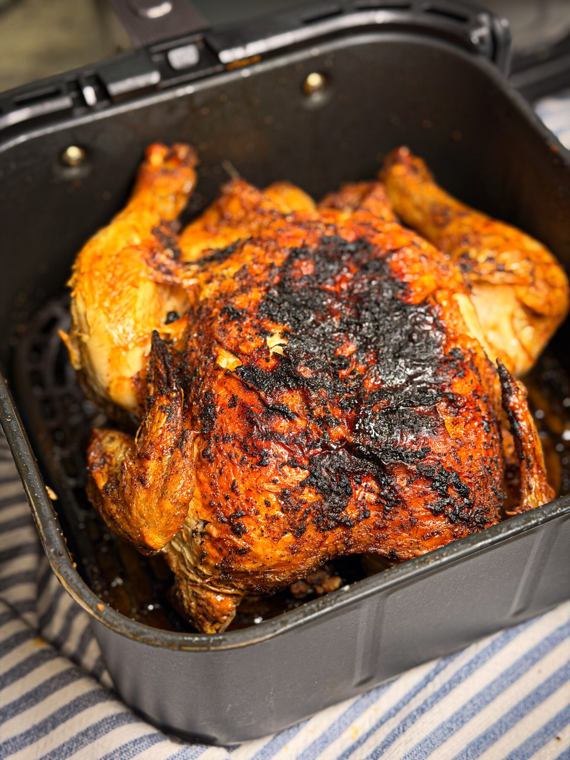 A whole roasted chicken with crispy, browned skin sits in an air fryer basket. The skin is seasoned and slightly charred in spots. The air fryer rests on a striped cloth surface.
