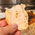 A hand with painted nails holds a slice of cooked, seasoned chicken breast in front of a whole roasted chicken.