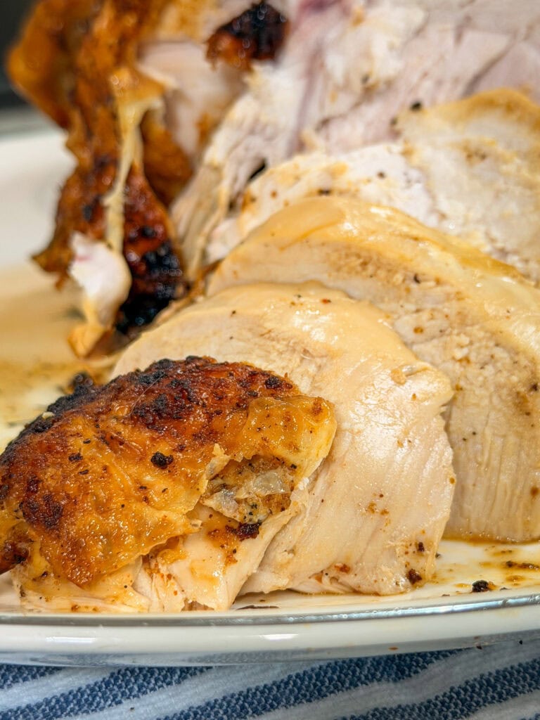 Sliced roasted turkey breast with crispy skin on a white plate, showing juicy, well-cooked meat. Some seasoning and juices are visible on the turkey and plate.