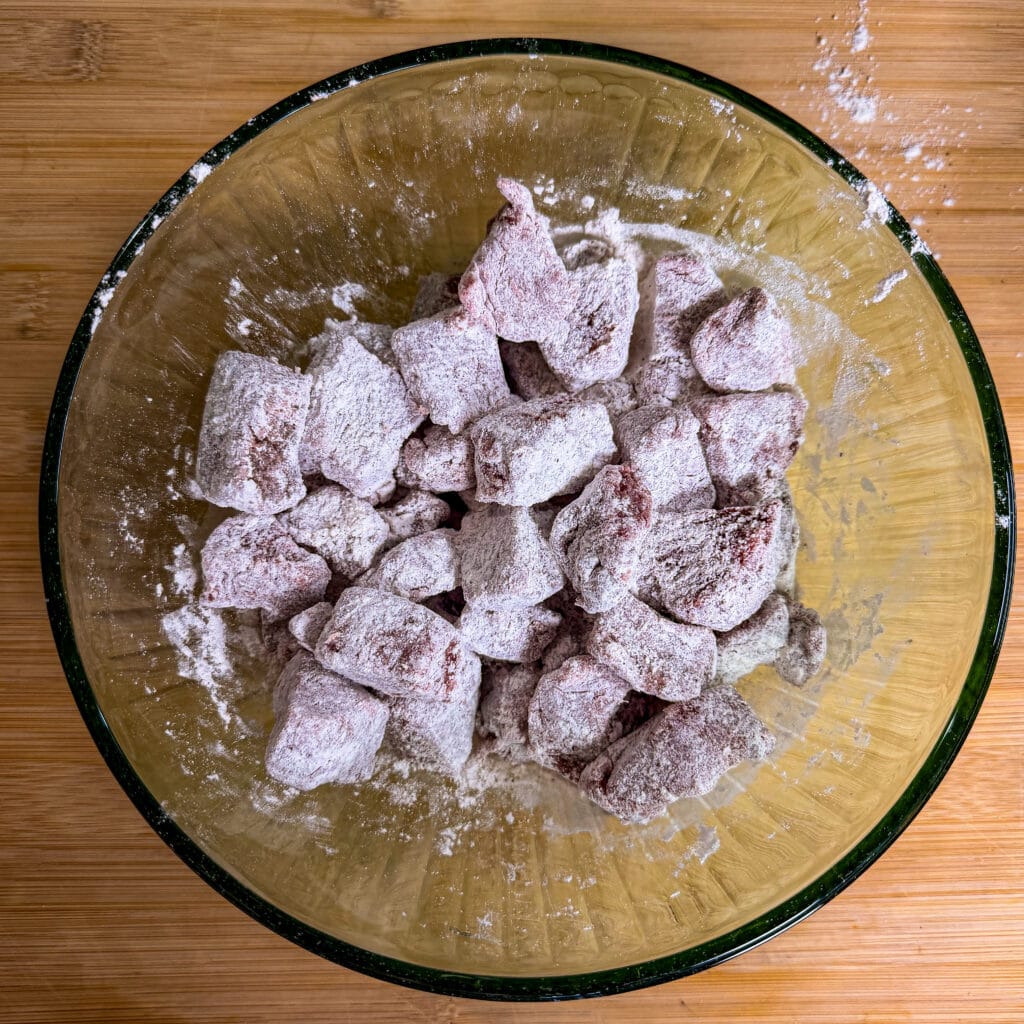 Chunks of raw meat coated in flour in a glass bowl, placed on a wooden surface.