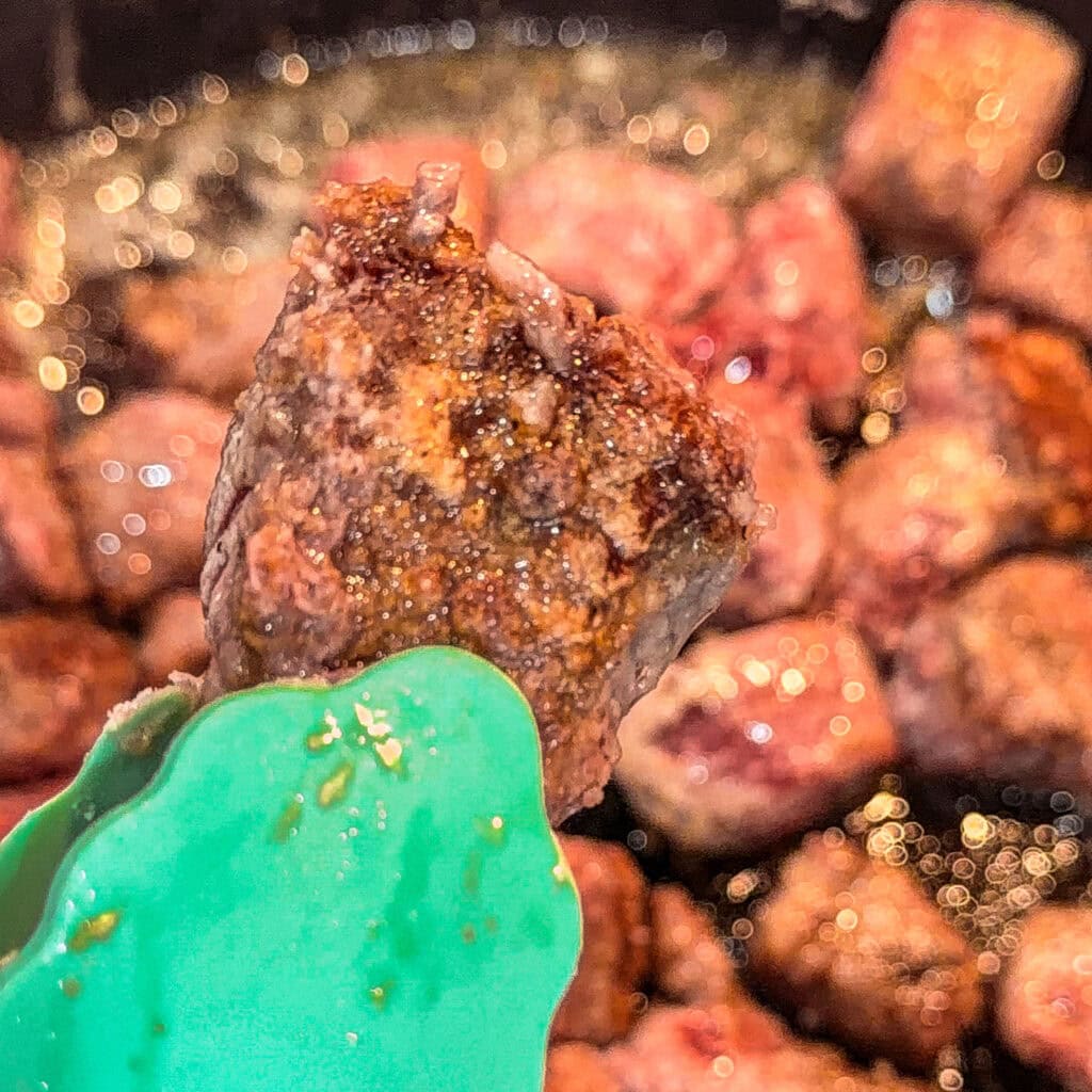 A close-up of green tongs holding a browned beef cube above a pan filled with more sizzling beef pieces.