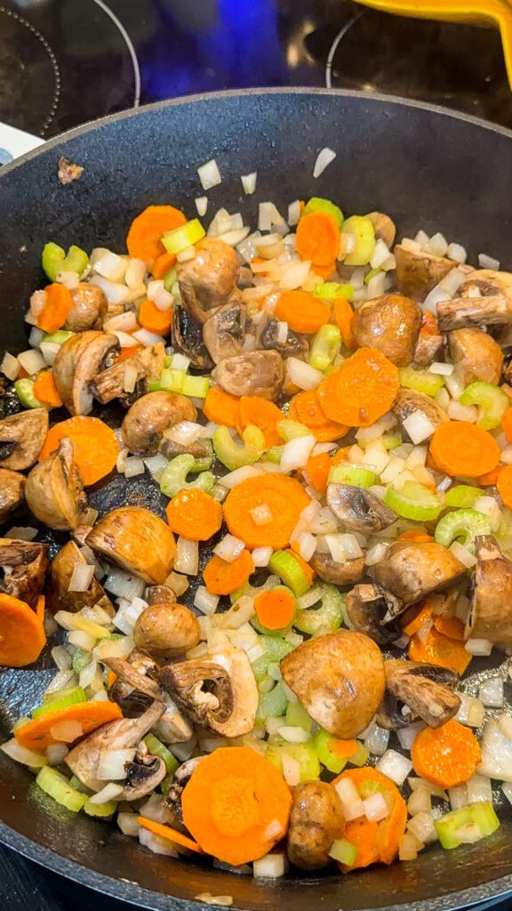 Chopped carrots, celery, onions, and mushrooms are being sautéed in a black frying pan on a stovetop.