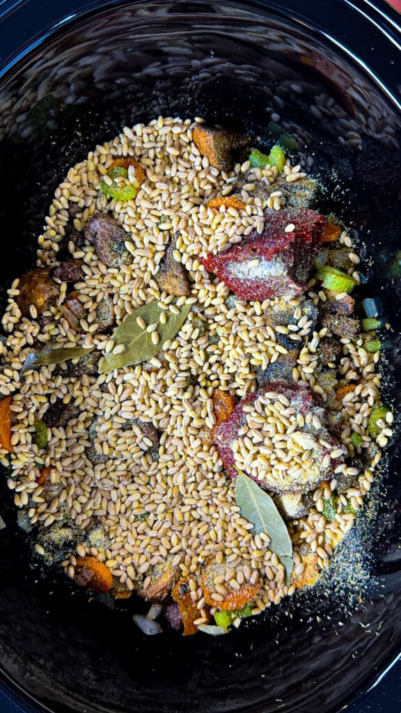 Uncooked soup ingredients in a slow cooker, including wheat berries, bay leaves, chopped vegetables, and seasonings.