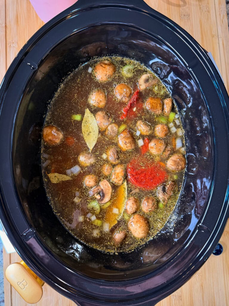 A slow cooker filled with broth, whole mushrooms, chopped onions, red bell peppers, bay leaves, and spices, sitting on a wooden surface.