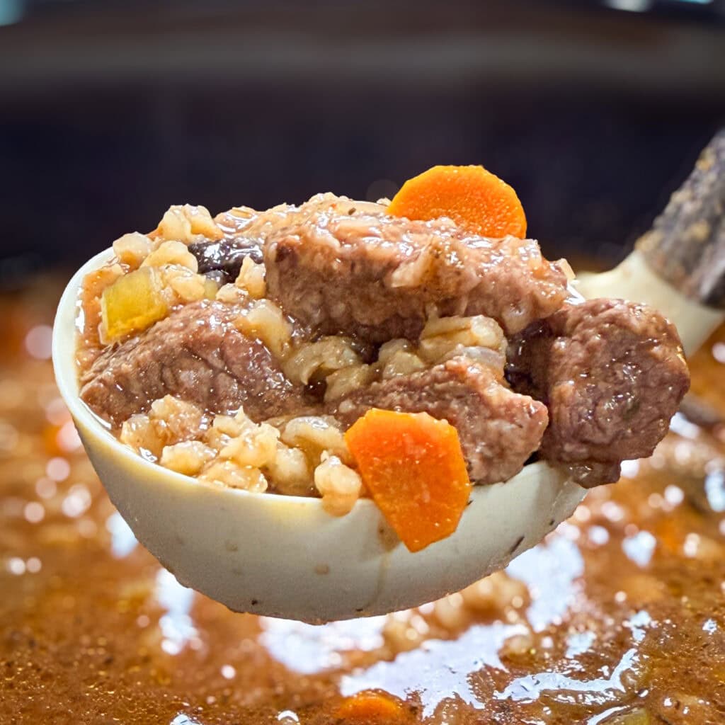 A ladle filled with hearty beef stew, showing chunks of beef, sliced carrots, barley, and potatoes in a rich brown broth.