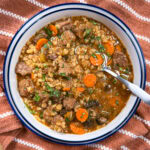 A bowl of beef and barley soup with chunks of beef, sliced carrots, celery, and parsley, placed on a rust-colored cloth with a white stripe pattern. A spoon rests inside the bowl.