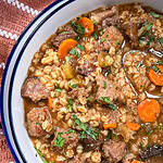 A close-up of beef and barley stew with chunks of beef, carrots, celery, barley, and fresh herbs in a white bowl on a rust-colored cloth.