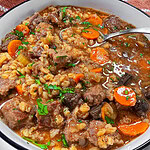 A bowl of beef and barley soup with chunks of beef, sliced carrots, celery, barley, and garnished with fresh parsley. A spoon rests in the bowl, revealing a hearty, savory broth.