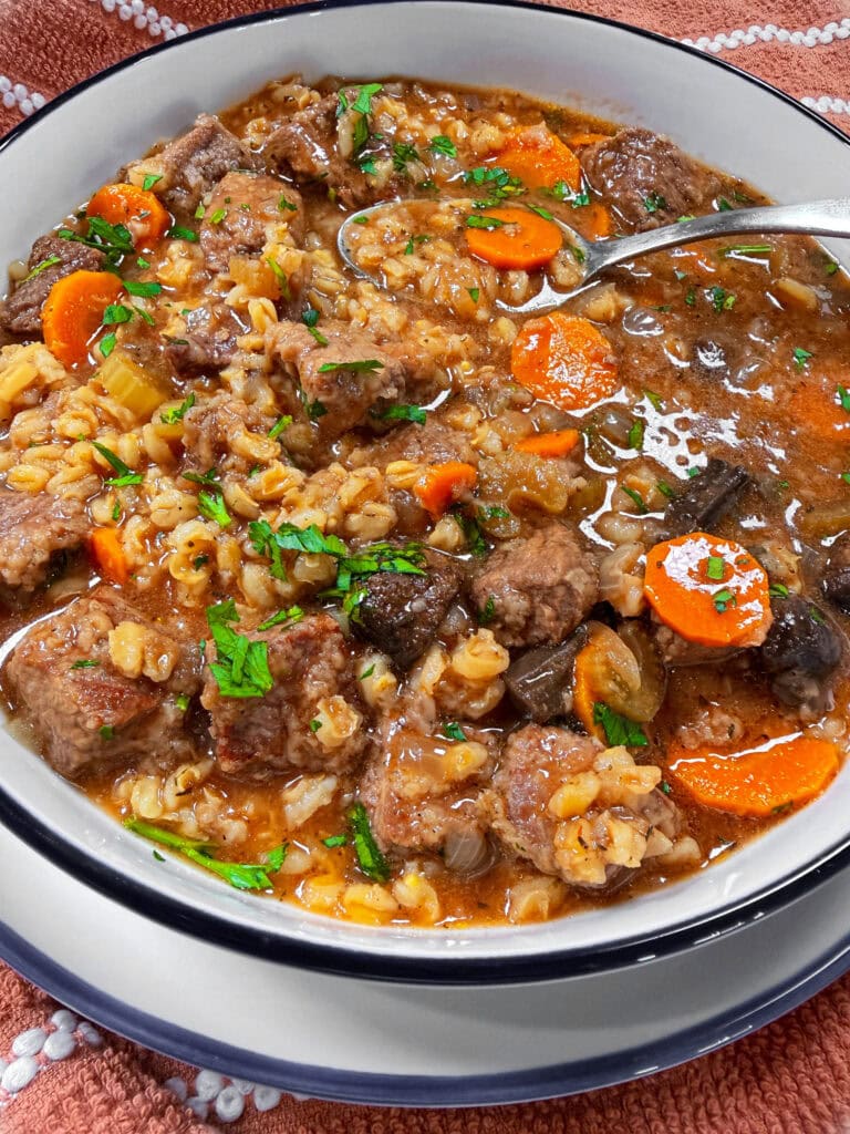 A bowl of beef and barley soup with chunks of beef, sliced carrots, celery, barley, and garnished with fresh parsley. A spoon rests in the bowl, revealing a hearty, savory broth.