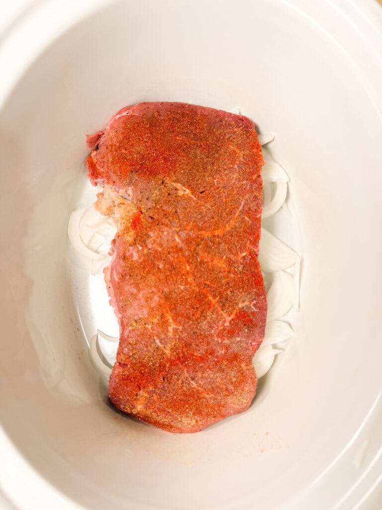 Raw seasoned beef sits on sliced onions in a white slow cooker, ready to be cooked.