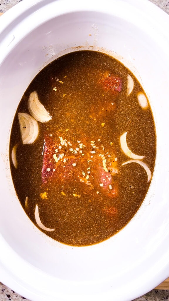 Raw beef, sliced onions, and spices submerged in dark broth inside a white slow cooker, ready to be cooked.