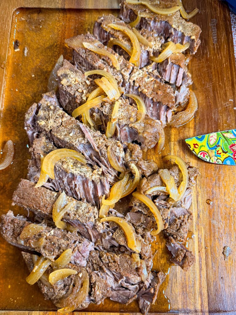 Sliced pot roast beef topped with cooked onions sits on a wooden cutting board. Pieces of beef are tender and juicy, with visible seasoning. A colorful knife is partially visible to the side.