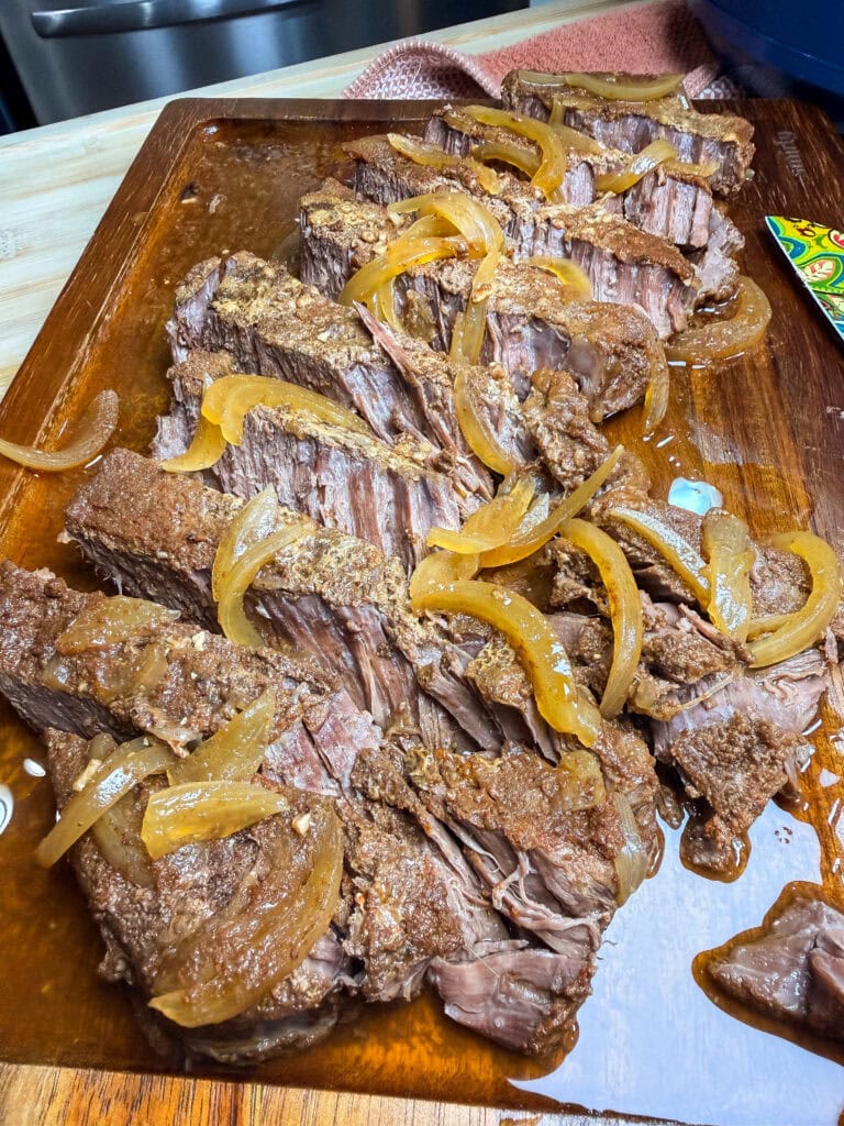 Sliced pot roast with onions and gravy on a wooden cutting board, showing tender beef pieces with visible seasonings and cooked onions on top.