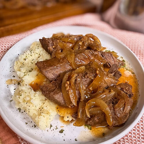 A plate with slices of roast beef topped with sautéed onions and gravy, served alongside mashed potatoes and garnished with herbs.