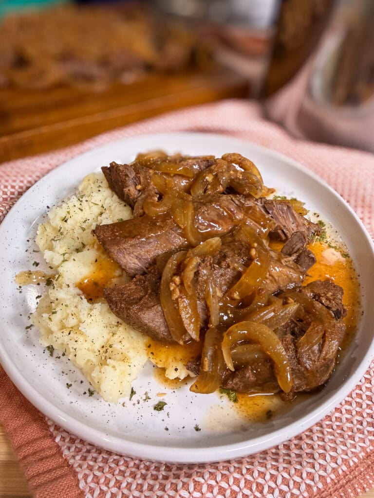 A plate with slices of roast beef topped with sautéed onions and gravy, served alongside mashed potatoes and garnished with herbs.