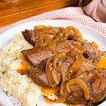 Plate of pot roast topped with caramelized onions and gravy, served with mashed potatoes. Food sits on a white plate with a pink textured napkin underneath.