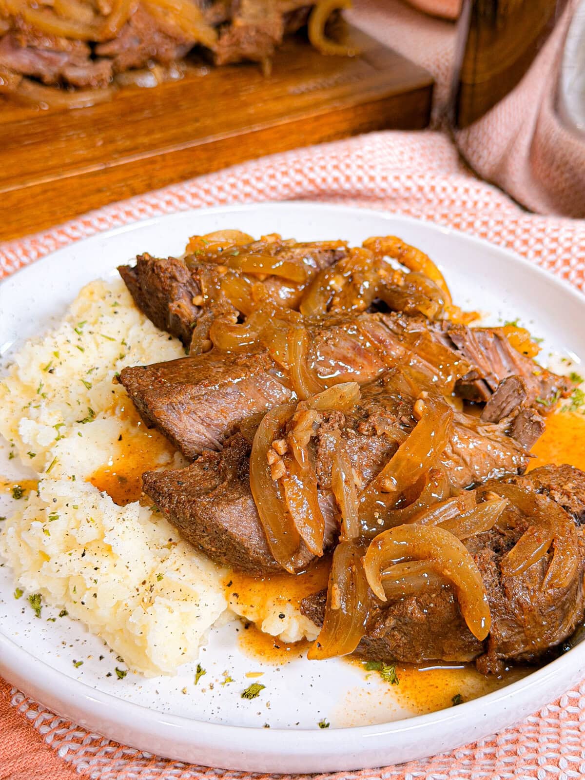 Plate of pot roast topped with caramelized onions and gravy, served with mashed potatoes. Food sits on a white plate with a pink textured napkin underneath.