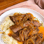 A plate of mashed potatoes topped with tender beef roast and caramelized onions in a rich brown sauce, garnished with herbs, on a white plate with a textured orange napkin nearby.