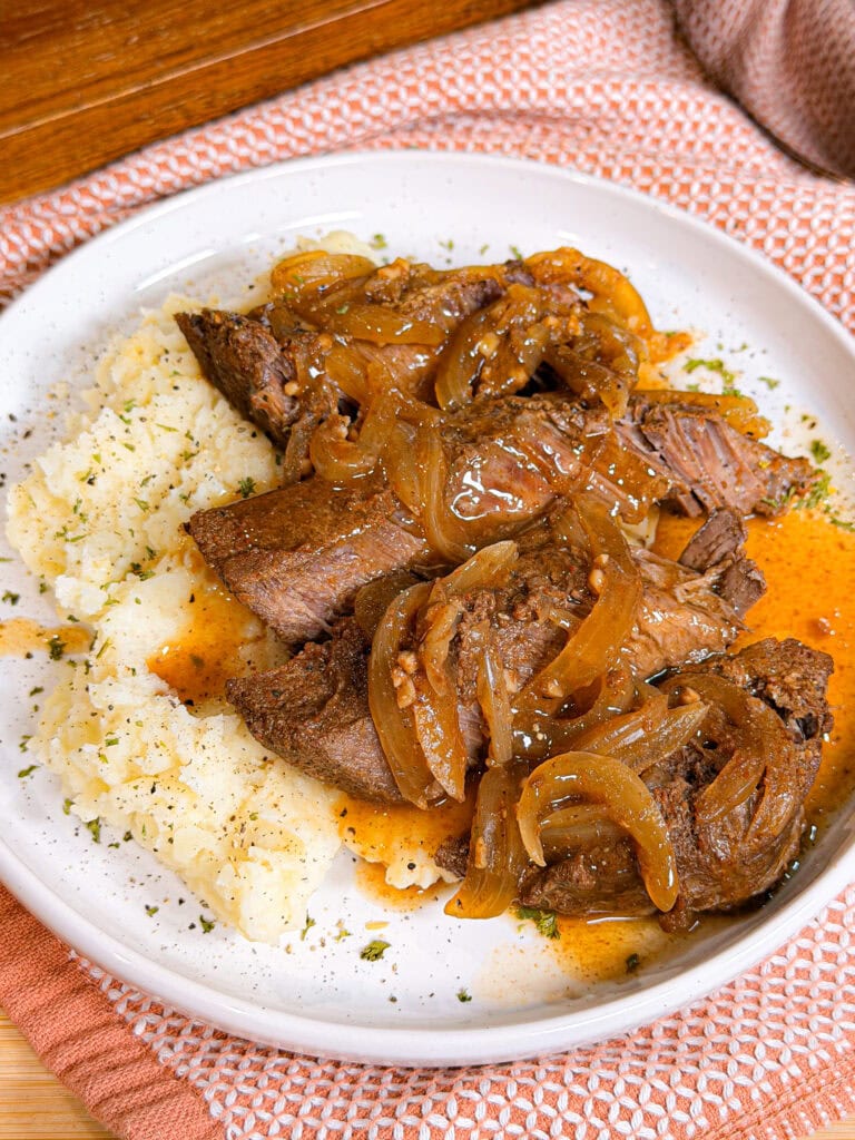 A plate of mashed potatoes topped with tender beef roast and caramelized onions in a rich brown sauce, garnished with herbs, on a white plate with a textured orange napkin nearby.